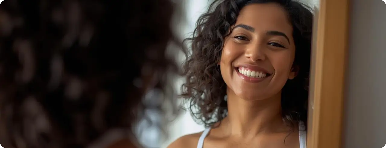 Woman with curly hair smiles at her reflection in a mirror, wearing a white tank top, in a brightly lit room.