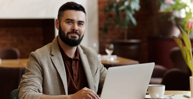 A man with a beard sits at a table in a café, working on a laptop with a cup of coffee nearby.