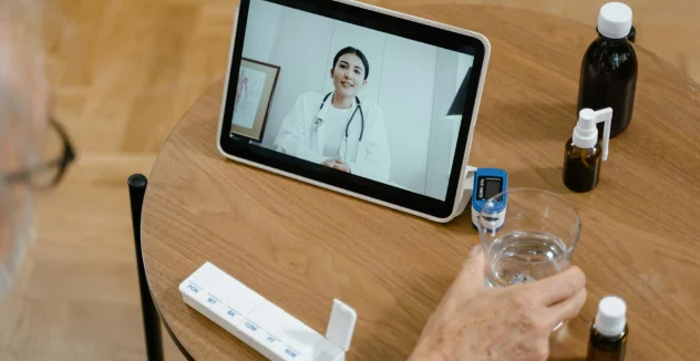 A person holds a glass of water while having a virtual consultation with a female doctor on a tablet. Medication and medical supplies are on the table.