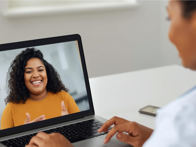 A person participates in a video call on a laptop, smiling and gesturing, while another person sits in front of the screen typing.