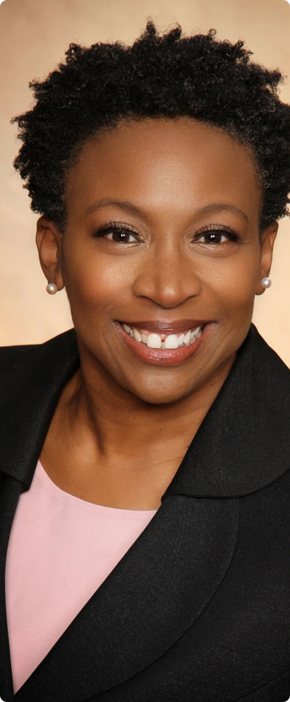 A person smiling, wearing a black blazer over a pink top, with short curly hair, pearl earrings, and a neutral background.