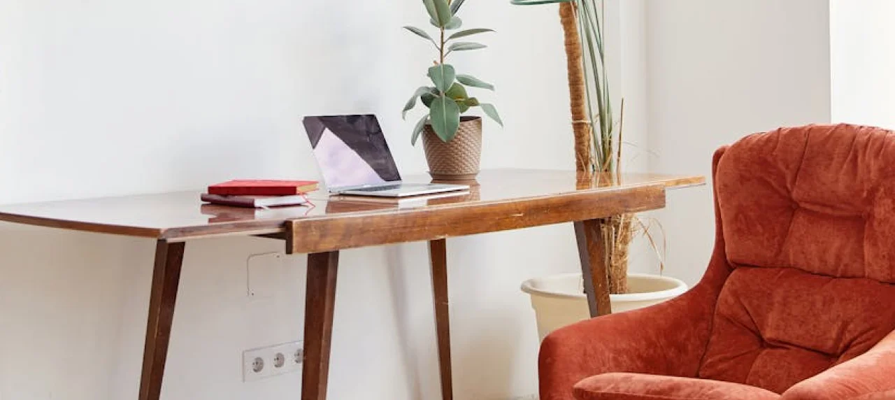 A wooden desk with a laptop, two books, a potted plant, and a red armchair in a bright room with white walls.