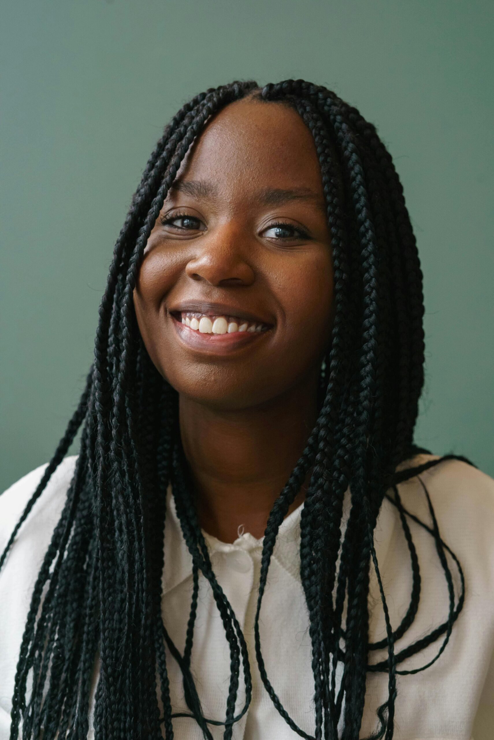 A young woman with long braids smiles warmly against a soft teal background, reflecting the welcoming, judgment-free care at Abundant Health & Wellness