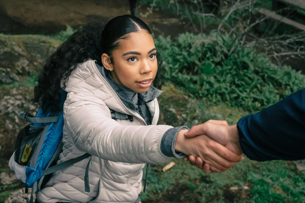 A woman with a backpack reaches up to accept a helping hand while climbing a rocky trail, symbolizing the compassionate support and partnership offered through behavioral health care at Abundant Health & Wellness