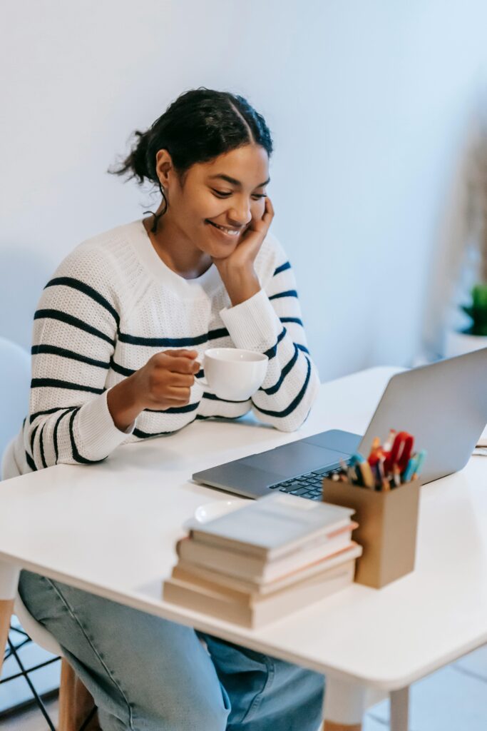 A woman smiles warmly while holding a coffee cup and viewing her laptop, representing a comfortable telehealth visit with Abundant Health & Wellness