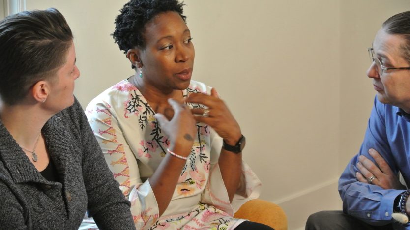 Three people sit together indoors having a serious conversation; the woman in the middle gestures while speaking to the others, who listen attentively.